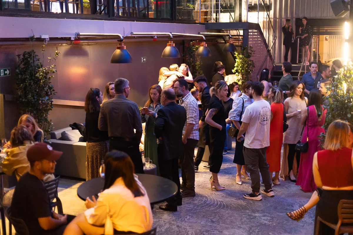 Ambiente iluminado e animado com dezenas de convidados interagindo durante um evento noturno. A decoração com plantas e luzes quentes cria um clima acolhedor e sofisticado para confraternizações empresariais e sociais.