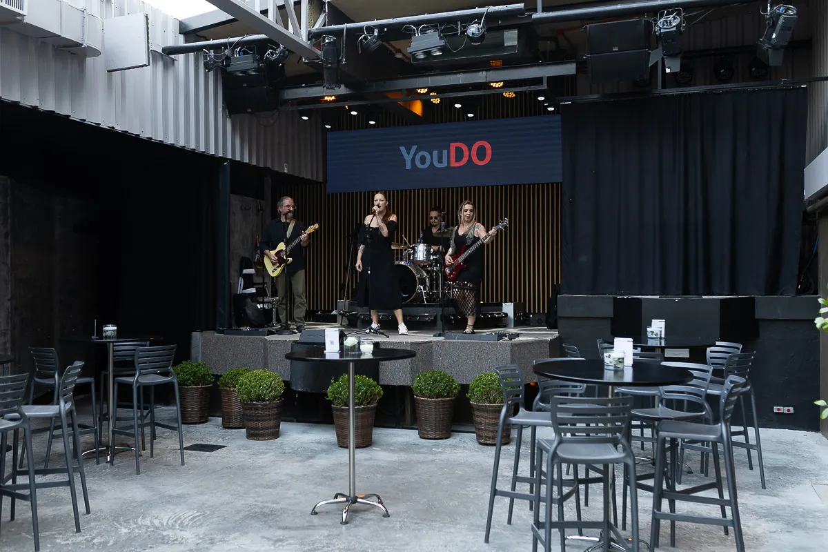 Palco com banda se apresentando ao vivo, rodeado por mesas e cadeiras. Ao fundo, a marca “YouDO” reforça a identidade do espaço, ideal para shows, festas e apresentações em um charmoso espaço para eventos em Curitiba.