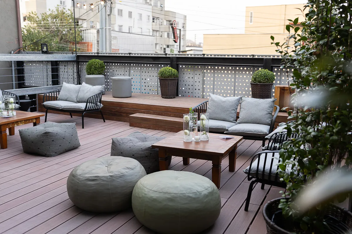 Área externa com deck de madeira, sofás modernos, pufes e mesa de centro com decoração floral. Um ambiente aconchegante ideal para confraternizações e eventos intimistas ao ar livre.