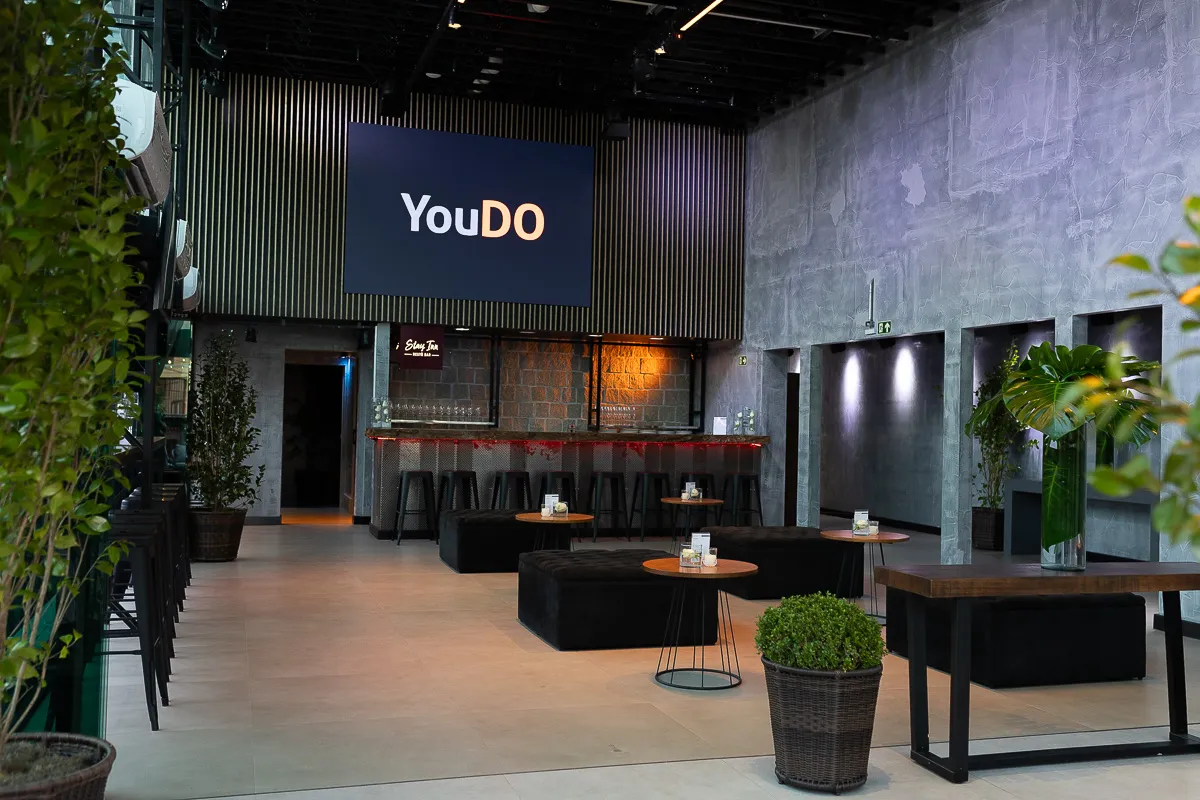 Ambiente interno sofisticado com decoração industrial, painel da marca "YouDO", bar de pedra com banquetas pretas e mesas baixas com pufes. Um espaço versátil para eventos sociais e corporativos.