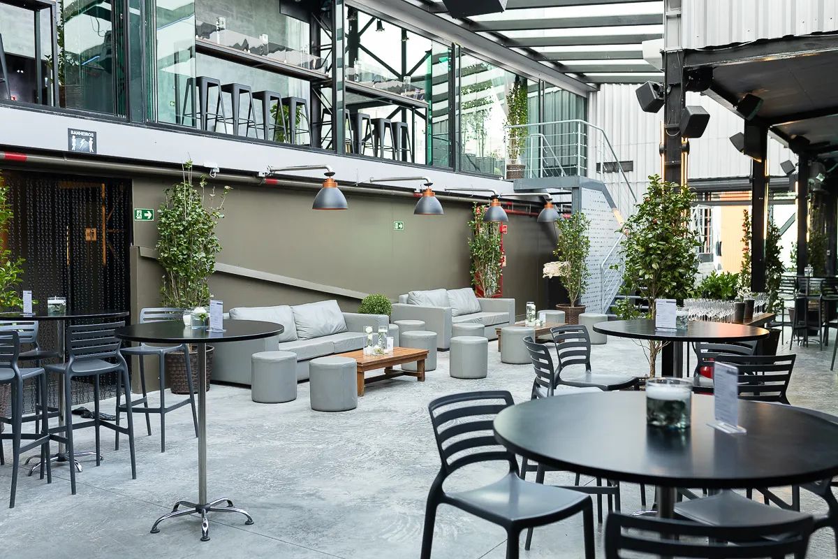 Área externa moderna com sofás, pufes e mesas redondas pretas, ideal para eventos sociais e confraternizações. O ambiente é decorado com plantas, iluminação industrial e mezanino com vista para todo o espaço.