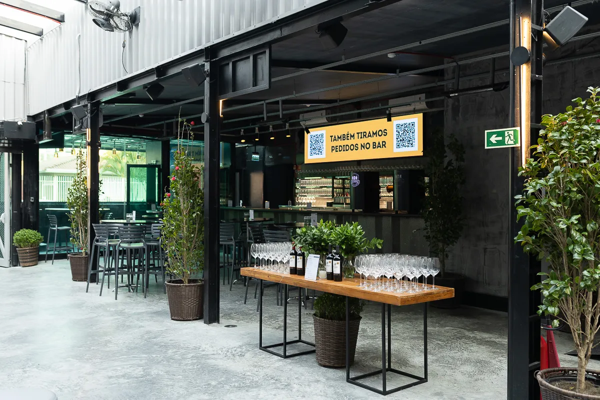 Área de bar moderna com estrutura metálica, banquetas altas e uma mesa com taças e vinhos pronta para recepção de convidados. Um painel sinaliza a praticidade dos pedidos via QR Code.