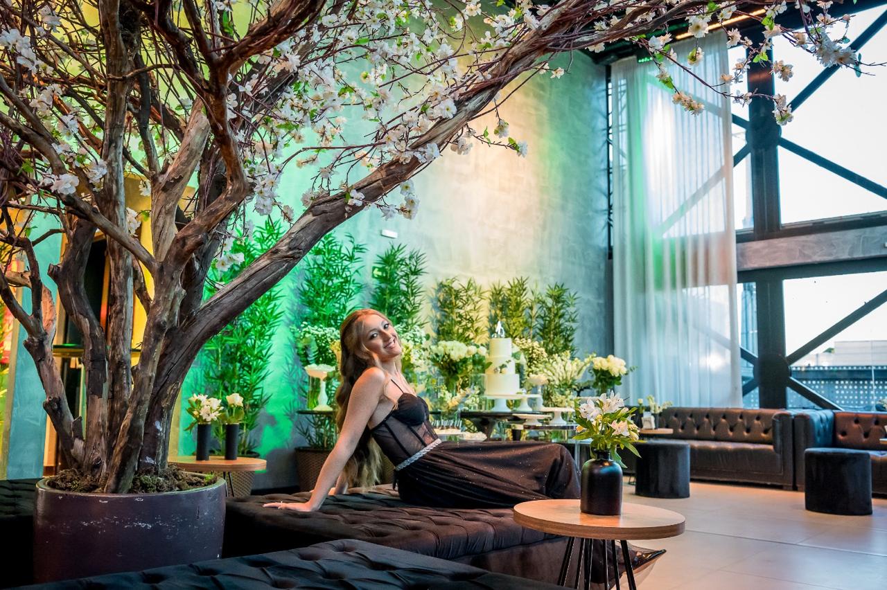Ambiente decorado com árvore de flores brancas, pufes, arranjos florais e uma convidada sorridente em traje de gala. Espaço charmoso perfeito para celebrações especiais como casamentos e aniversários.