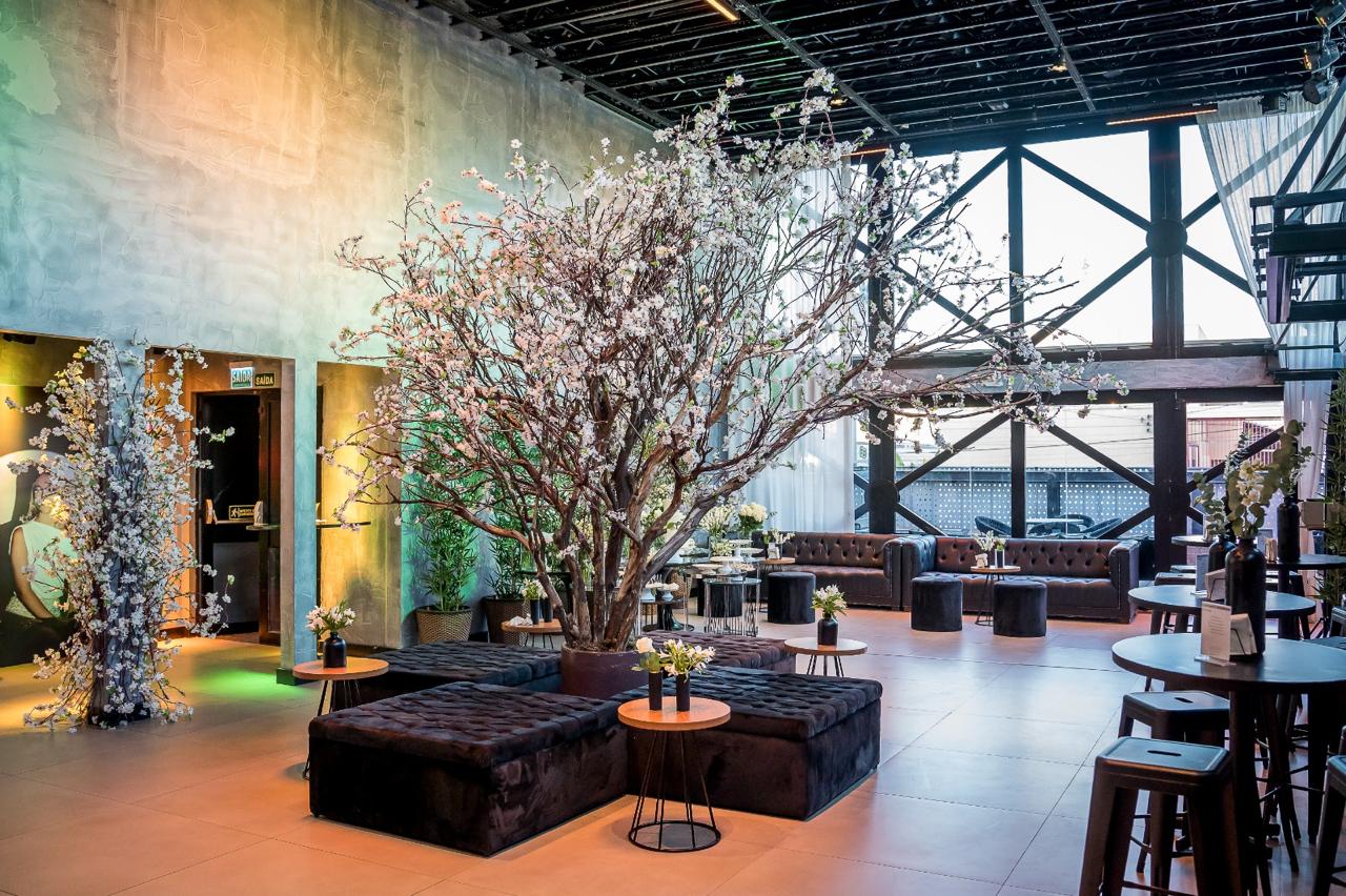 Vista ampla de um espaço para eventos em Curitiba, decorado com árvore de flores no centro, sofás, pufes e mesas redondas. Ambientes integrados com iluminação cênica e detalhes naturais.