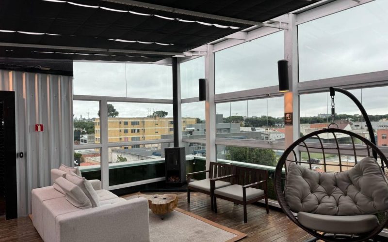 Área de estar envidraçada de uma cobertura mobiliada no Rebouças, com sofá em L, mesa de centro rústica e lareira moderna, ideal para quem busca cobertura para alugar em Curitiba com estrutura aconchegante e vista urbana.