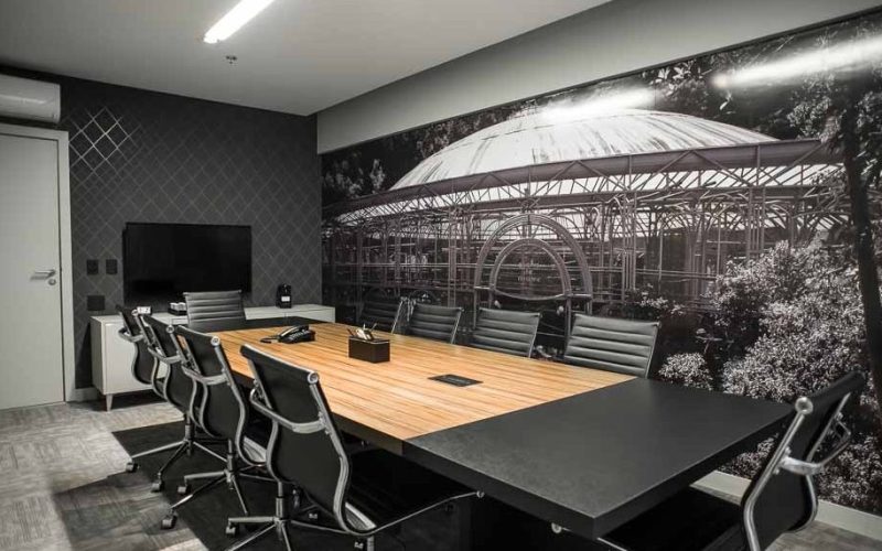 Sala de reuniões com mesa retangular de madeira clara, várias cadeiras pretas com encosto vazado e parede decorativa com imagem em preto e branco da Ópera de Arame. Um ambiente elegante e executivo.
